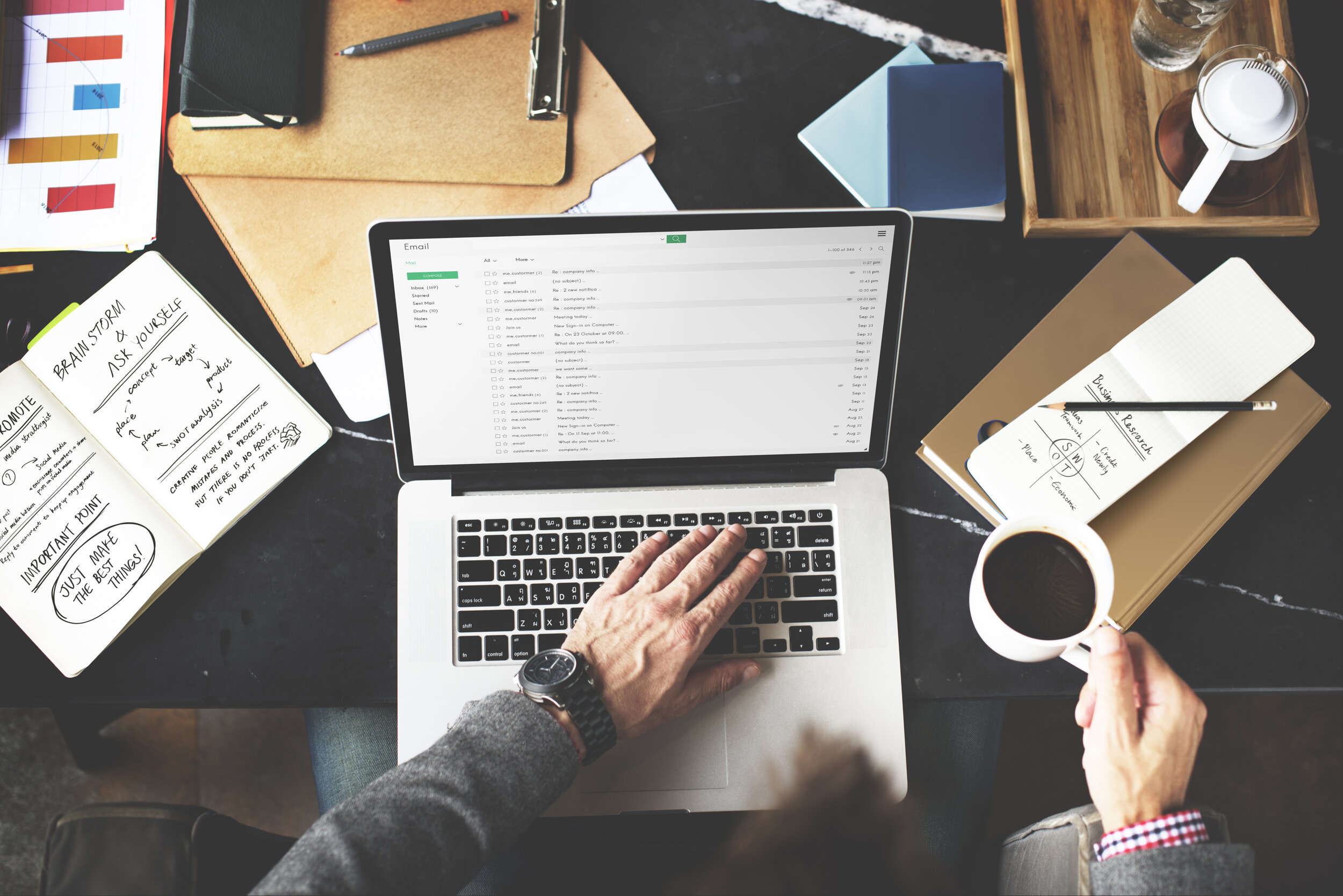 An overview shot of a person sitting writing emails on an Apple laptop and a cluttered desk.