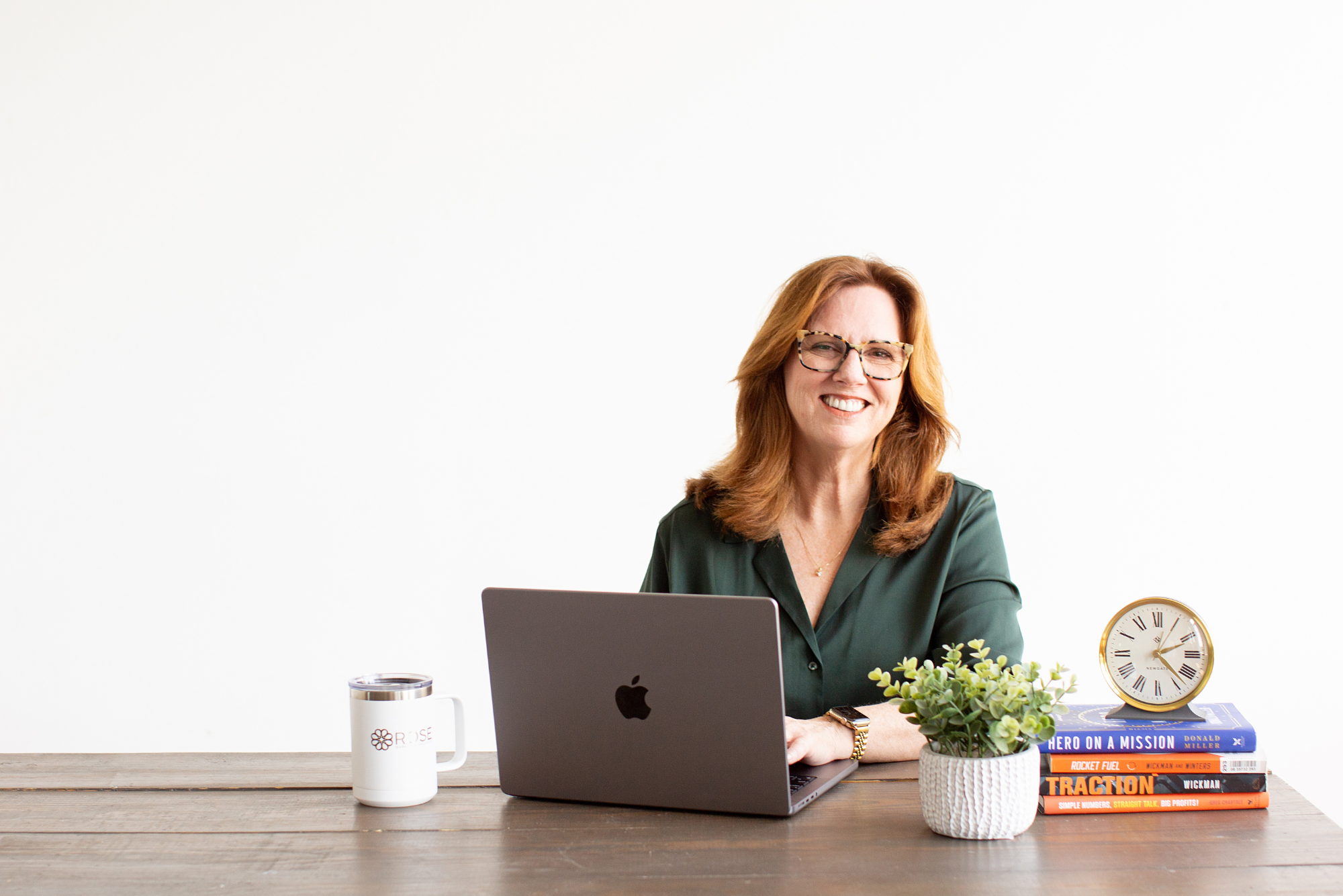 Ruth Ann smiles while working on her laptop with a stack of business books and a potted plant next to her.